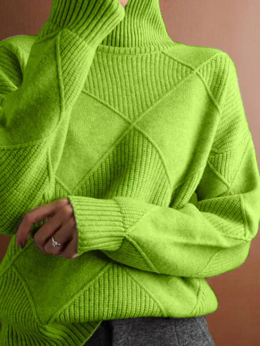 A close-up of a person wearing a bright lime green ribbed turtleneck sweater with diamond-shaped knit patterns on the front and sleeves, paired with gray bottoms against a solid warm brown background.