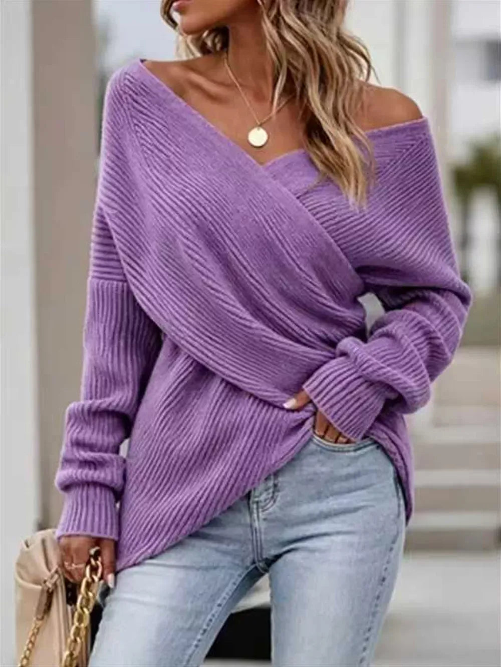 A woman wearing a lavender purple off-the-shoulder long-sleeve knit sweater with a wrap-style crossover front, paired with light-wash blue jeans.