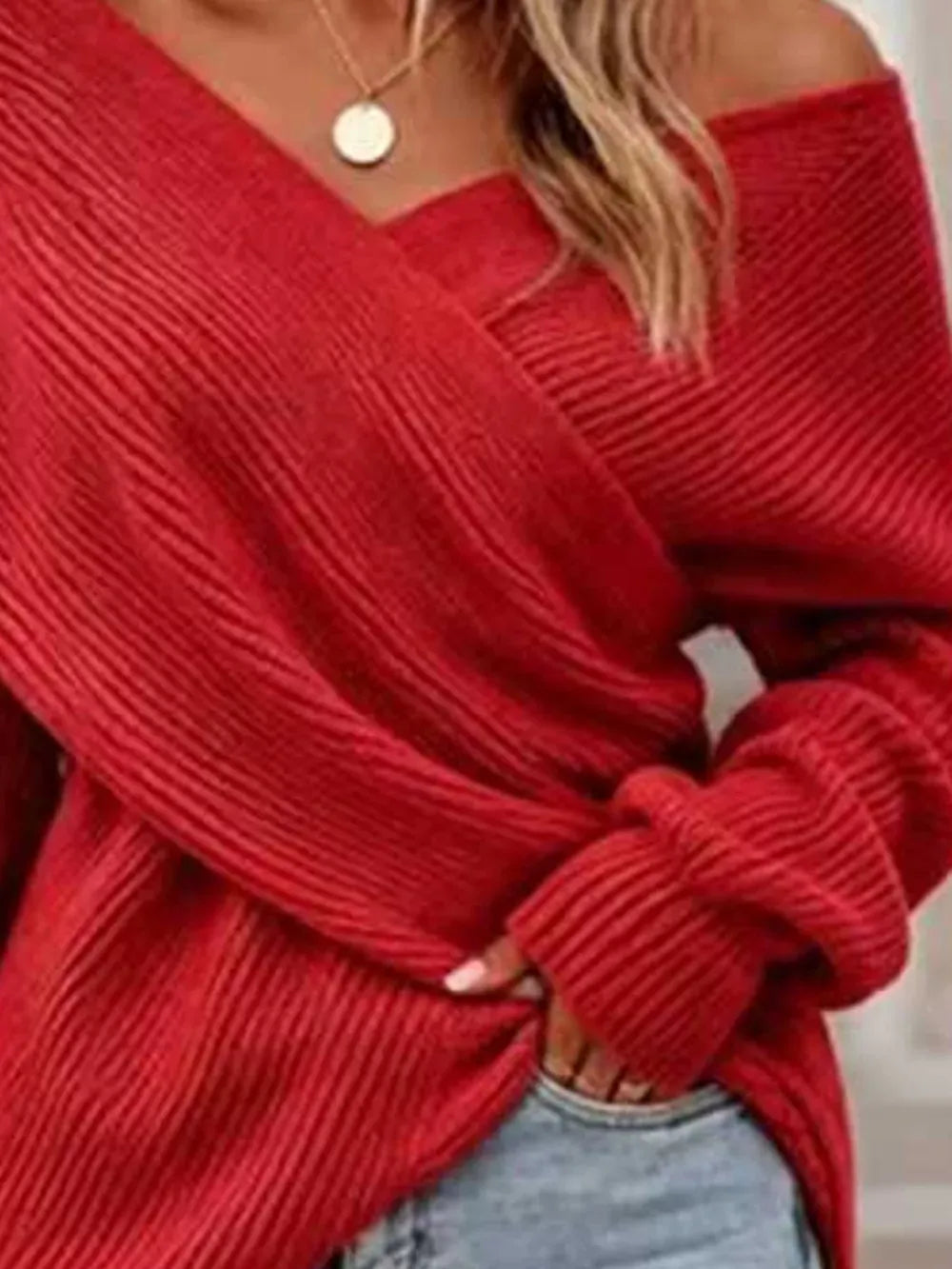 A close-up of a woman wearing a red off-the-shoulder ribbed knit sweater with light blue jeans and a gold pendant necklace.
