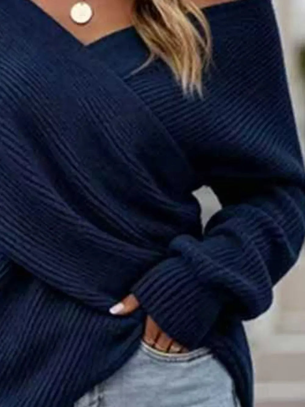 A close-up of a person wearing a navy blue ribbed knit sweater with an off-the-shoulder wrap-style neckline and light gray jeans.
